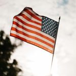 Low angle of crop faceless person holding spiky stick with flag of USA under cloudy sky and tree in back lit on blurred background